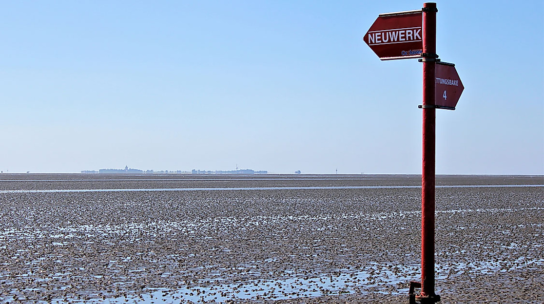 Wegweiser im Watt vor Neuwerk bei Ebbe, zeigt Richtung Cuxhaven und Insel, im Hintergrund Wattenmeer und Horizont.