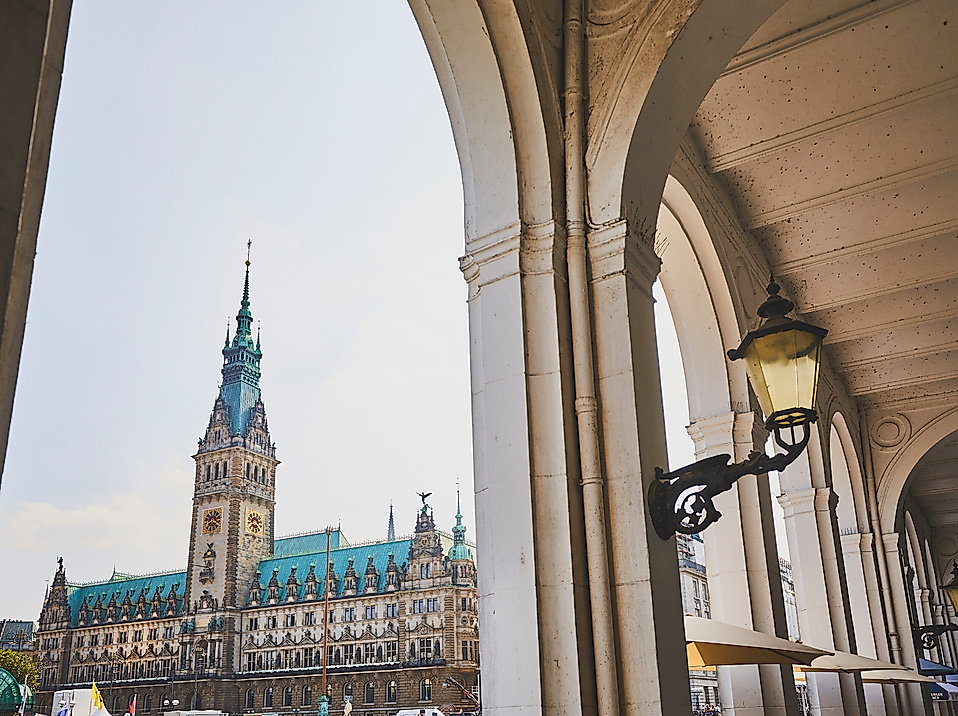 Blick durch Alsterarkaden auf das Hamburger Rathaus mit grünem Turm und klassizistischer Fassade.