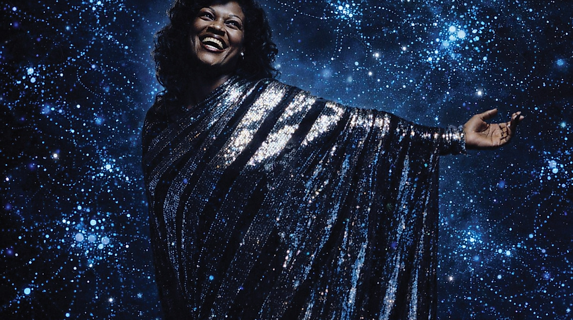 Eine Frau steht in einem silber-glitzernden Kleid vor dem Hintergrund einer dunkelblauen Fläche, auf der viele Lichter sind.