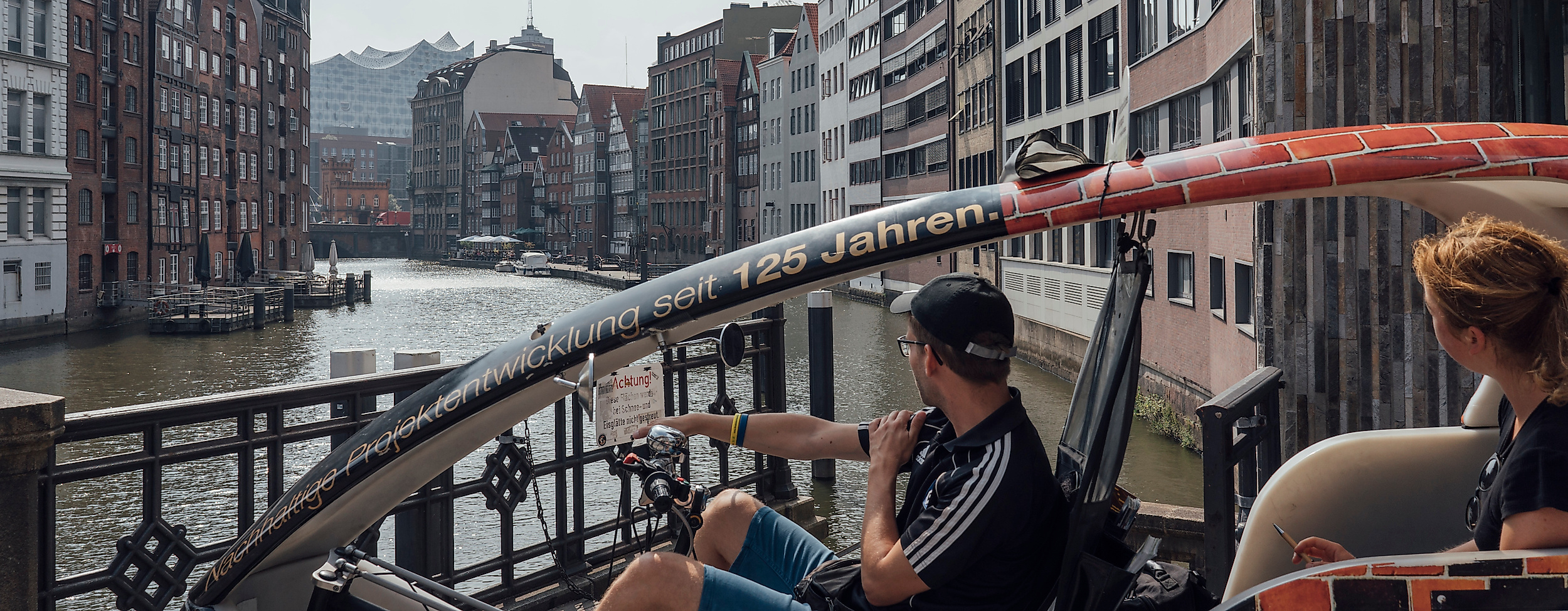 Zwei Personen fahren mit einer Rikscha durch die Hamburger Speicherstadt entlang eines Fleets.