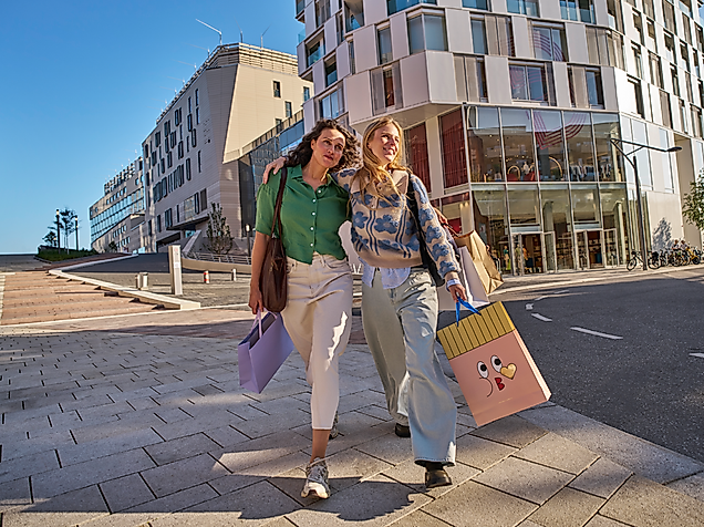 Two women stroll in the sunshine along the Hamburg Alster in front of Westfield Shopping Centre