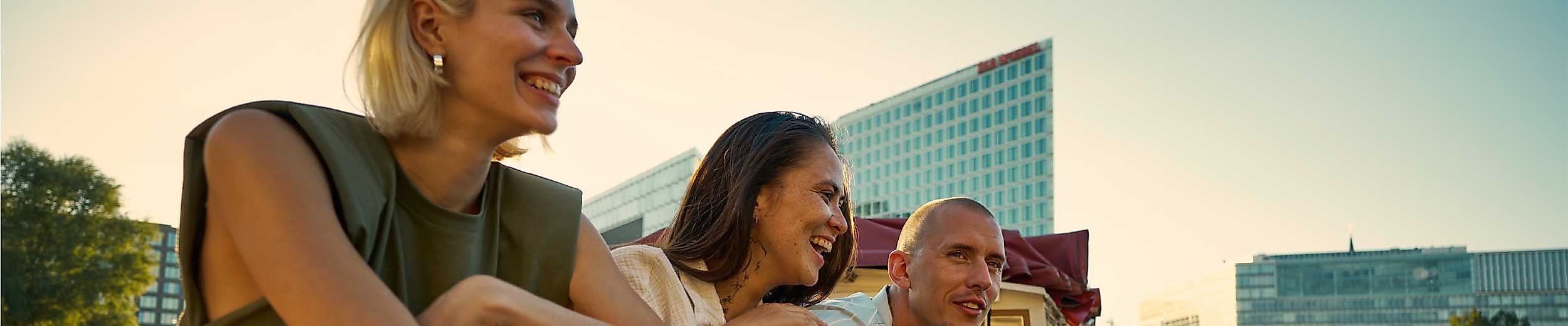 Drei Personen stehen fröhlich an der Reling eines Ausflugsschiffs auf einem Gewässer in Hamburg. Sie schauen lachend in die Ferne. Im Hintergrund ist moderne Architektur zu sehen, vermutlich die Hafencity. Die Sonne steht tief und taucht die Szene in warmes Licht.