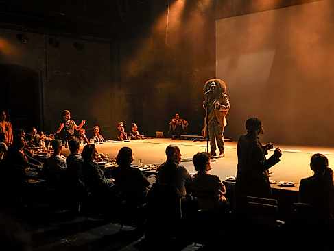 A moody theatre scene: people sit at a long table filled with food while a person on stage speaks or sings into a microphone. Warm light, intimate atmosphere, sense of community.
