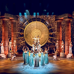 Szenenbild Disneys HERCULES: Person in weißem Gewand im Mittelpunkt des Olympus, umgeben von Tänzerinnen in Türkis vor Säulen und Lichtern.