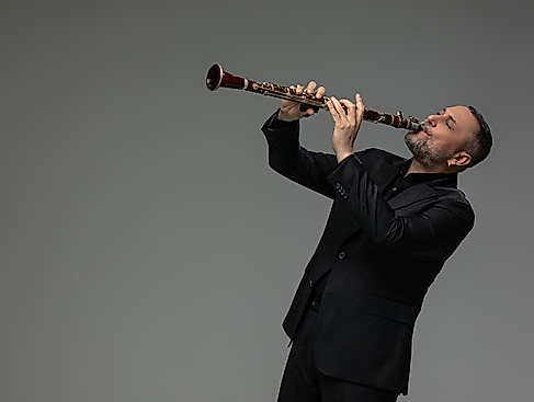 A man in a black suit and short gray beard plays the clarinet in front of a gray background. He bends slightly backwards.