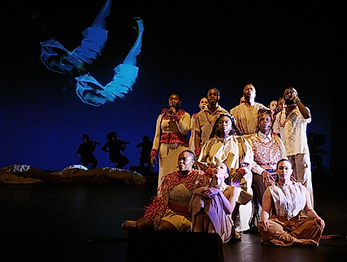 A group of performers in beige and red costumes sit and stand in formation on stage. In the background, silhouettes of musicians and a projection of a floating body are visible.