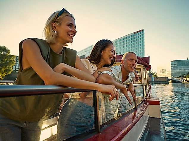 Drei Personen stehen fröhlich an der Reling eines Ausflugsschiffs auf einem Gewässer in Hamburg. Sie schauen lachend in die Ferne. Im Hintergrund ist moderne Architektur zu sehen, vermutlich die Hafencity. Die Sonne steht tief und taucht die Szene in warmes Licht.