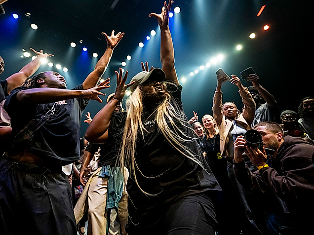 A dynamic scene in which a crowd of people stand around a dancer, cheering and cheering her on with gestures. The dancer raises her arms upwards, spreading her fingers. At the bottom right, someone photographs the dancer from close up.