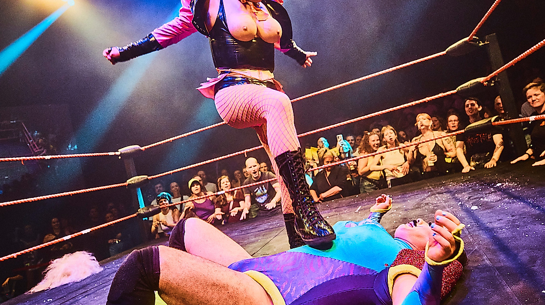 Eine Wrestling-Szene: eine Person liegt auf dem Boden, eine andere Person steht über ihr und hat ihren Fuß auf dem Brustkorb der anderen Person abgestellt.