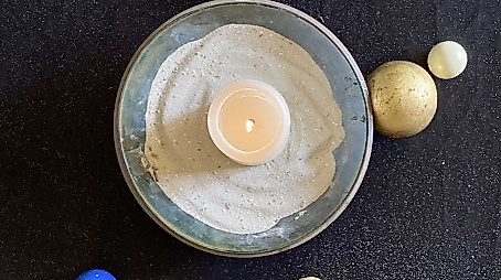 Kerze in einer Sandschale rundherum ist angeordnet ein Kreuz und verschiedenfarbige Perlen