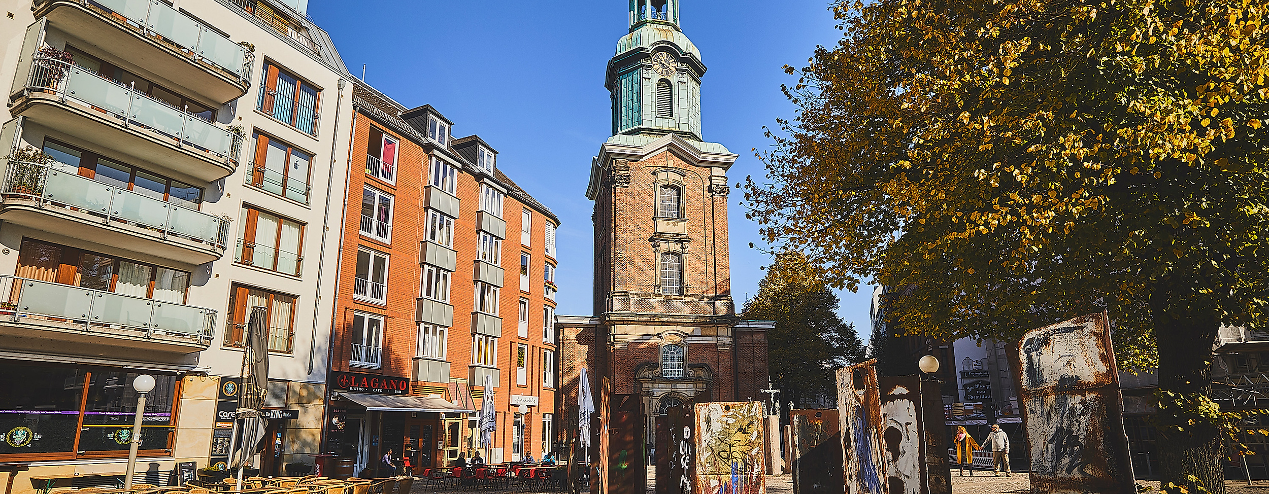 Belebter Platz an der St.-Georg-Kirche in Hamburg mit Außengastronomie, Kunstobjekten und herbstlicher Stimmung