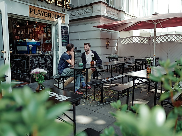 Zwei Personen sitzen im Außenbereich von Playground Coffee, umgeben von Pflanzen und roten Sonnenschirmen