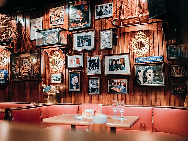 Gemütliche Kneipe mit rot gepolsterter Sitzbank, Holztischen und Wänden voller gerahmter Fotos
