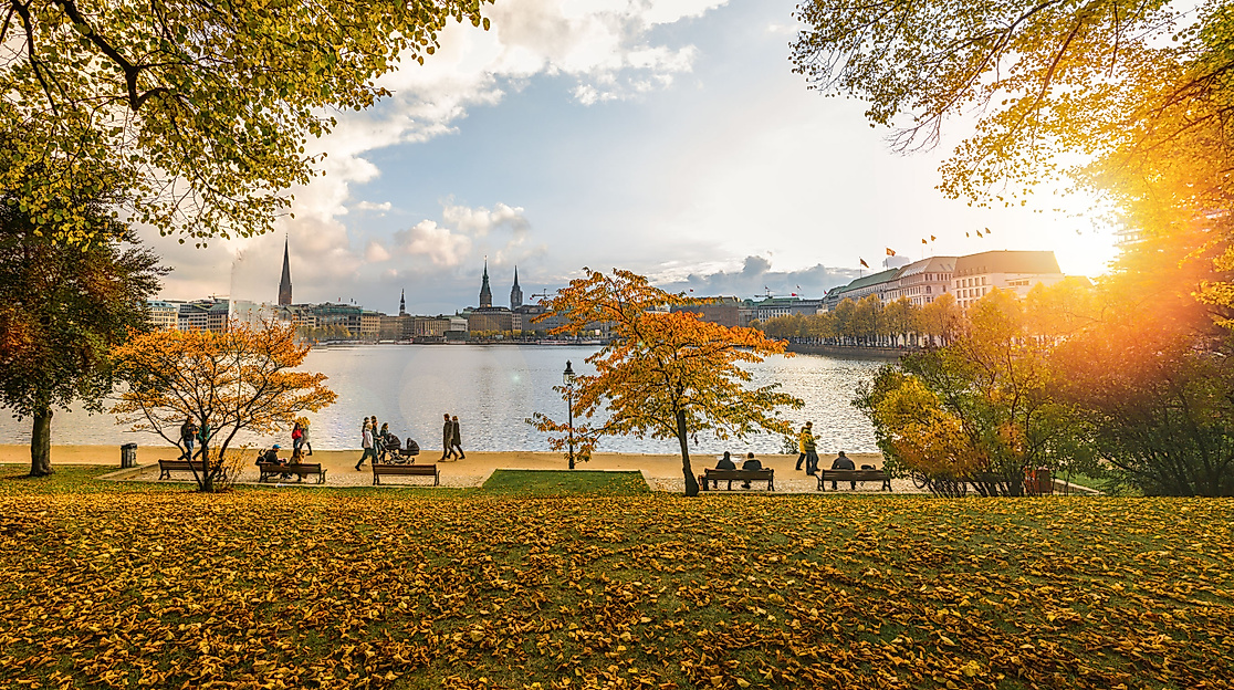 Herbstlicher Spazierweg an der Außenalster mit goldenem Laub und Blick auf Hamburgs Altstadt im Sonnenlicht