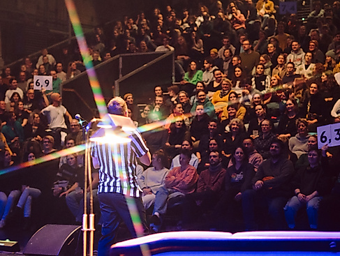 A person wearing a black and white striped T-shirt stands in front of a large audience. Two boards displaying scores are held up in the audience.