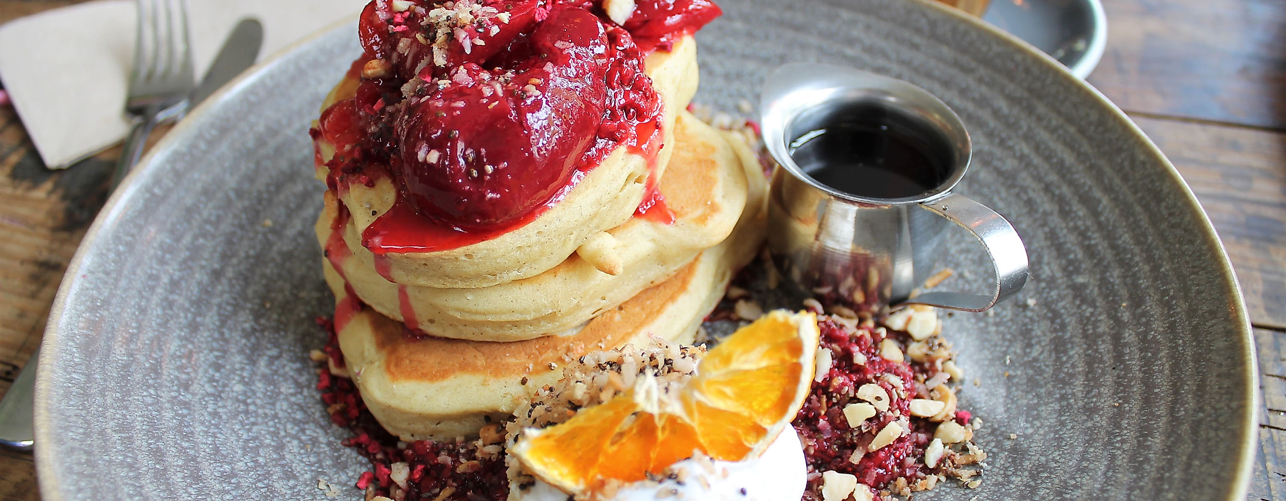 Pancake-Frühstück mit Beeren, Kokoscreme, Orangenscheibe und Ahornsirup auf grauem Teller angerichtet