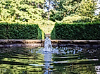 Wasser sprudelt aus einem Brunnenbecken, eingefasst von Hecken und Bäumen in einer ruhigen Parkanlage.
