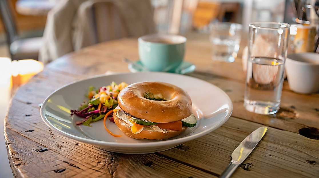 Frühstücksbagel auf weißem Teller, dazu Tasse und Glas auf Holztisch