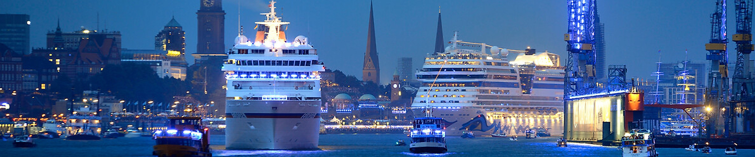 Blick auf den Hamburger Hafen bei Nacht während der Hamburg Cruise Days: Zwei beleuchtete Kreuzfahrtschiffe fahren in Richtung Landungsbrücken, begleitet von kleineren Hafenbooten und umgeben von blau-violett illuminierten Kränen. Im Hintergrund leuchten die Silhouetten von Michel, Kirchtürmen und der Hamburger Skyline.