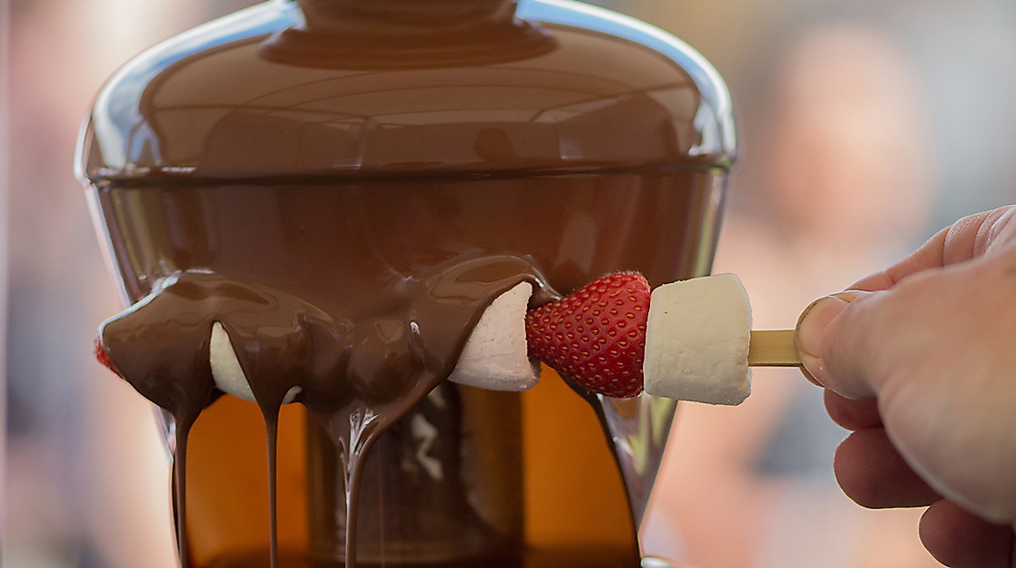 Schokoladenbrunnen mit fließender Kuvertüre, darin wird Obstspieß mit Erdbeeren und Bananen getaucht.
