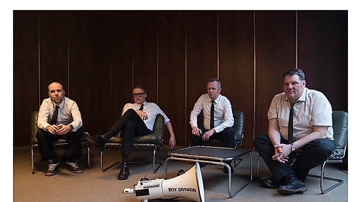 The four members of Boy Division all wear a white shirt and a narrow black tie; they sit casually in a '60s-style conference room, with a white megaphone in front of them, labeled with the band's name.