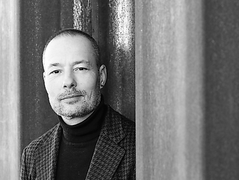 Black-and-white photograph of a male-read person with short-cropped hair, a black earring, a beard, and dressed in a plaid jacket.