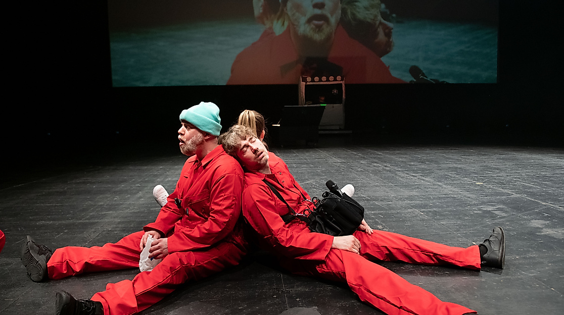 Three performers in red jumpsuits sit back-to-back on a stage with their eyes closed. Behind them, a close-up of them is projected onto the wall.