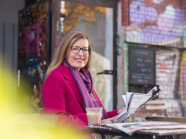 Lächelnde Frau mit Zeitschrift und Getränk im Straßencafé vor bunt besprühter Wand im Hamburger Schanzenviertel.