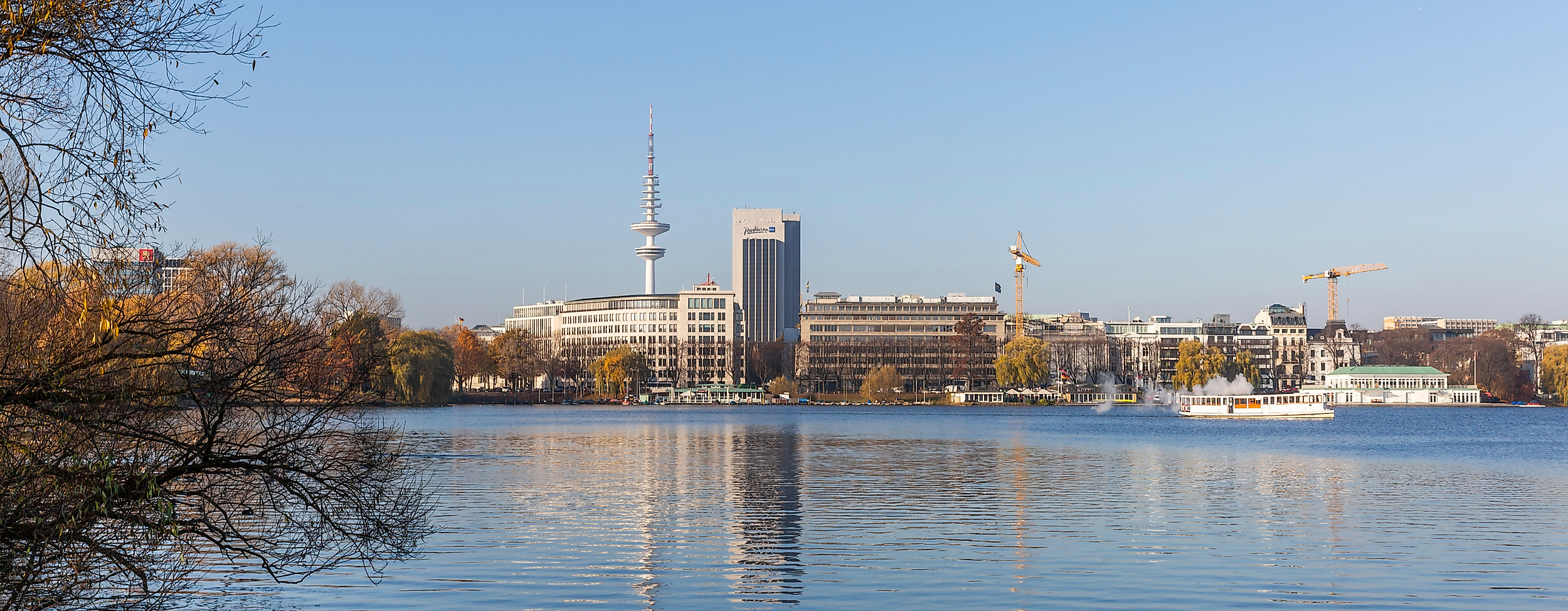 Blick über die Außenalster in Hamburg mit Uferbäumen, Segelboot und markanter Stadtsilhouette im Sonnenlicht.