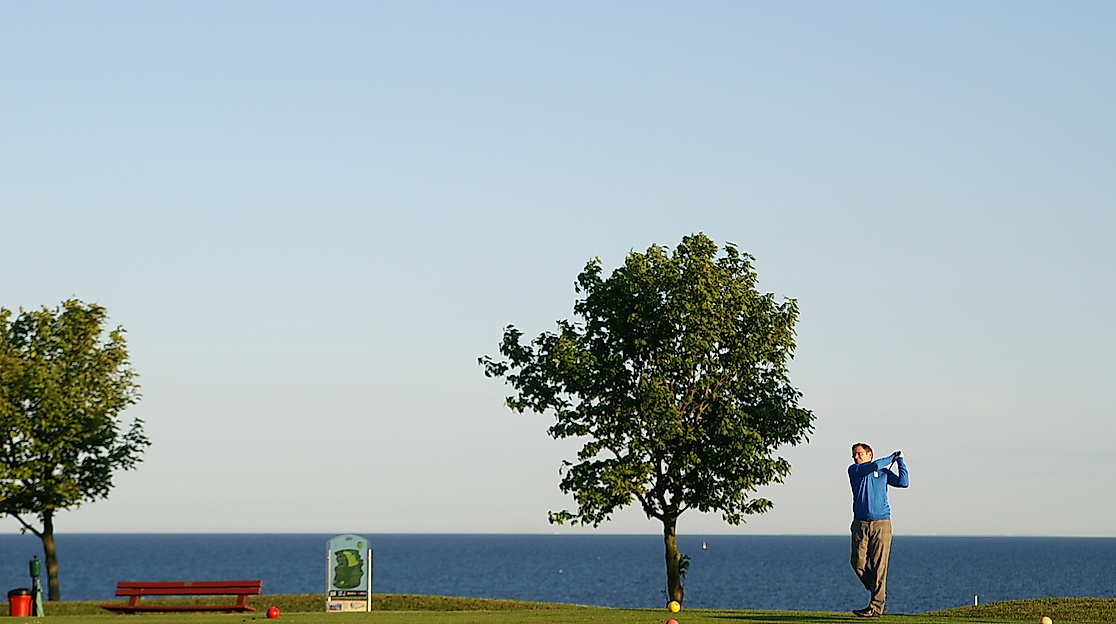Golfer schlägt am Golfpark Fehmarn mit Blick auf die Ostsee ab, umgeben von Bäumen und Küstenlandschaft