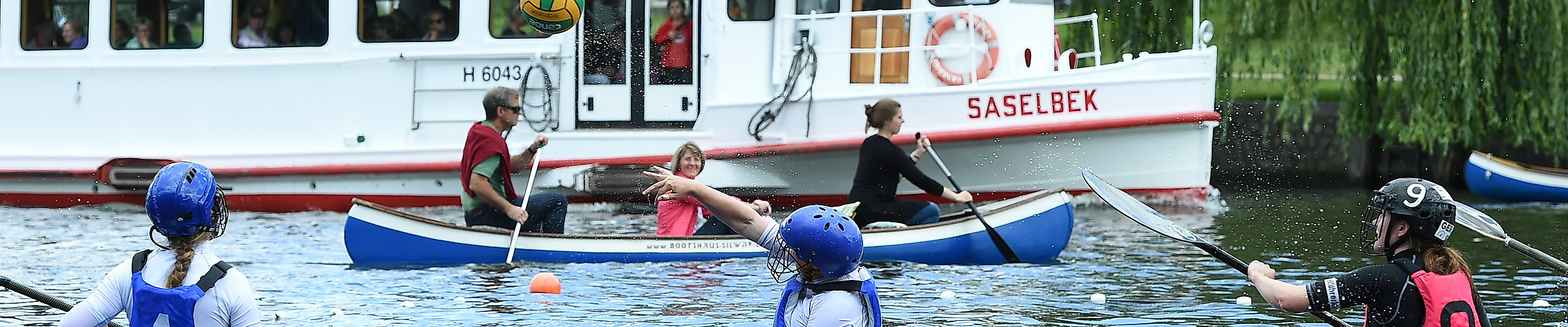 Kanupolo-Spieler:innen im Wettkampf auf der Außenalster, im Hintergrund ein weißes Ausflugsschiff