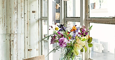 Vase mit frischen, bunt gemischten Blumen auf Holztisch vor Fensterfront in hellem Kreativraum