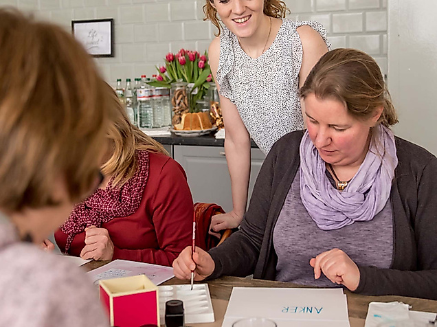 Workshop-Leiterin unterstützt Teilnehmende beim kreativen Lettering an einem Tisch mit Übungsblättern und Tinte