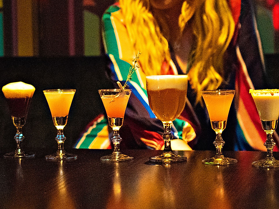 Sechs bunte Cocktails auf dunkler Theke, dahinter Person in bunter Kleidung vor Clubkulisse