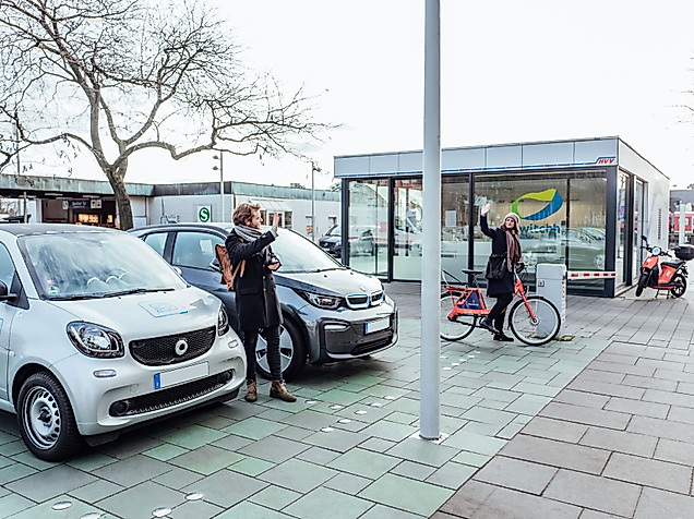 Zwei Mietwägen mit davorstehendem Mann, welcher einer Frau auf einem Stadtrad winkt. 