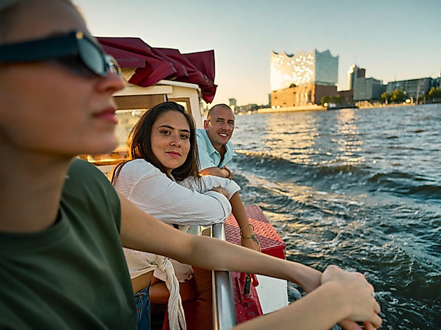 Drei junge Menschen auf einem Boot auf der Elbe. Im Hintergrund ist die Elbphilharmonie.