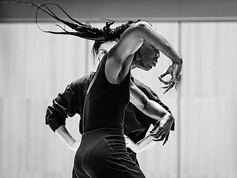 A performer is seen from the side, moving her arms expressively. In the background, a second person can be seen watching her.