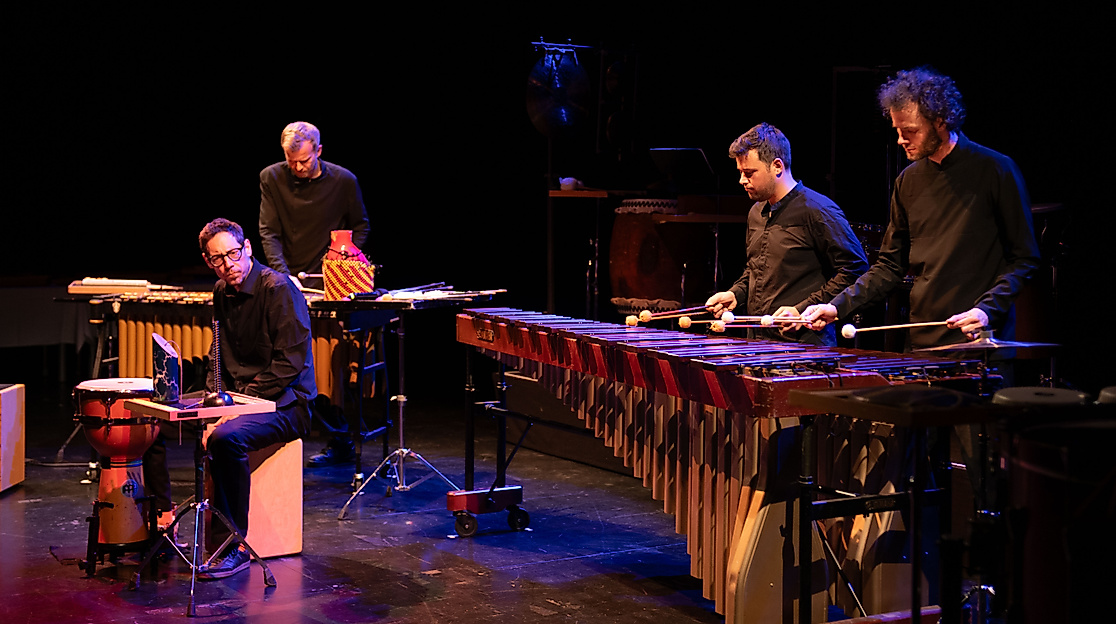 Three musicians play various percussion instruments. They are all dressed in black and bathed in atmospheric stage lighting.