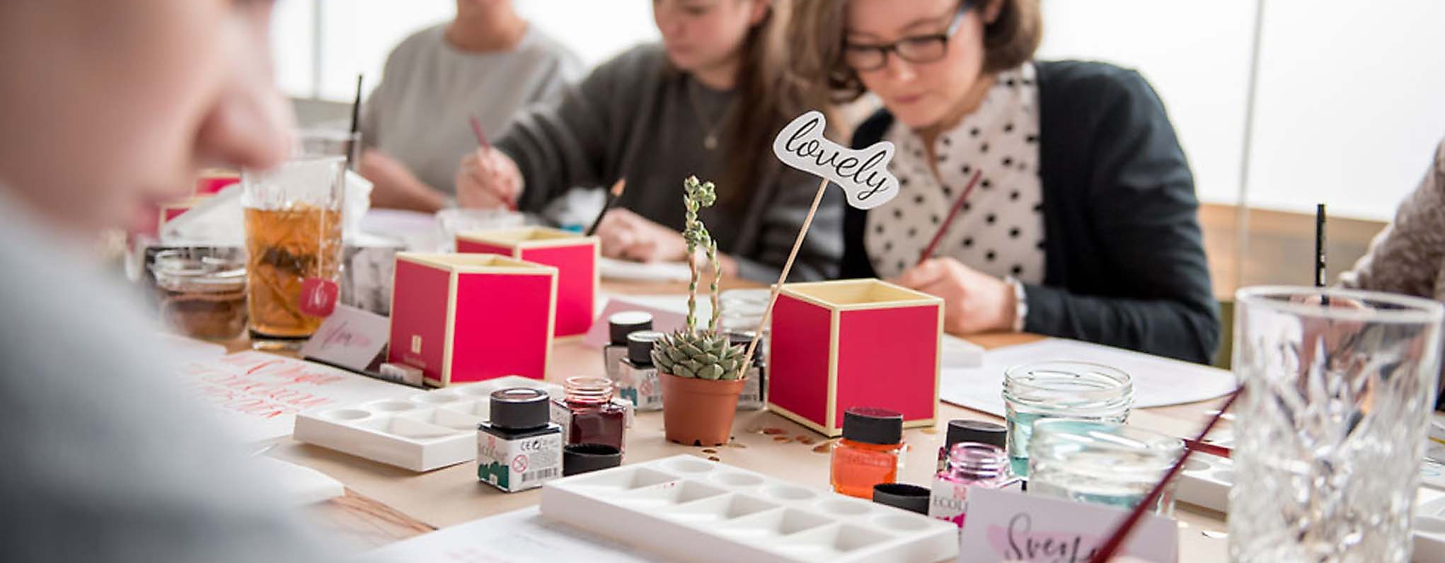 Mehrere Personen arbeiten konzentriert an einem langen Tisch mit Lettering-Vorlagen, Pinseln und Tinte.