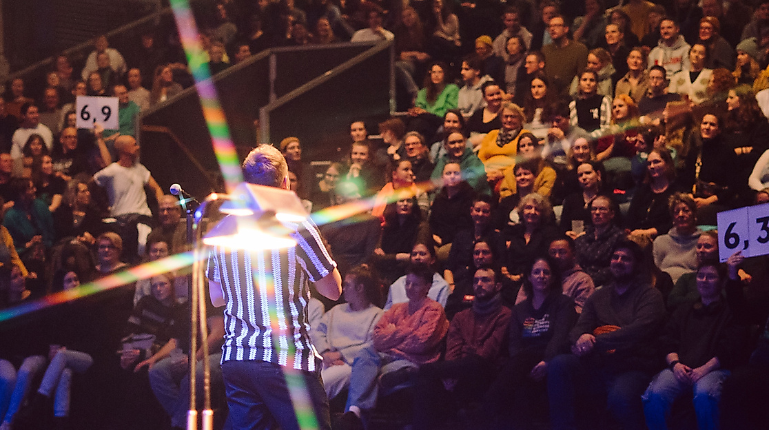 A person wearing a black and white striped T-shirt stands in front of a large audience. Two boards displaying scores are held up in the audience.