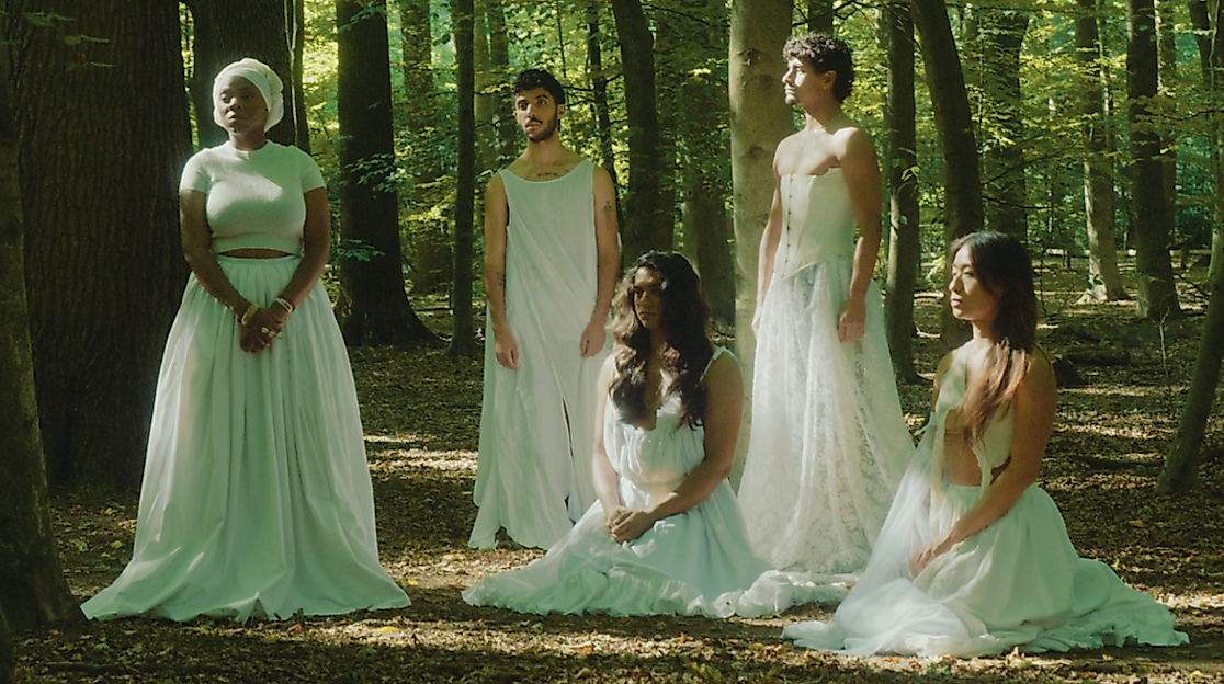 Fünf Performer*inne in weißen langen Kleidern posieren im Wald. Drei von ihnen stehen, zwei sitzen auf dem Boden. Keine*r schaut in die Kamera.