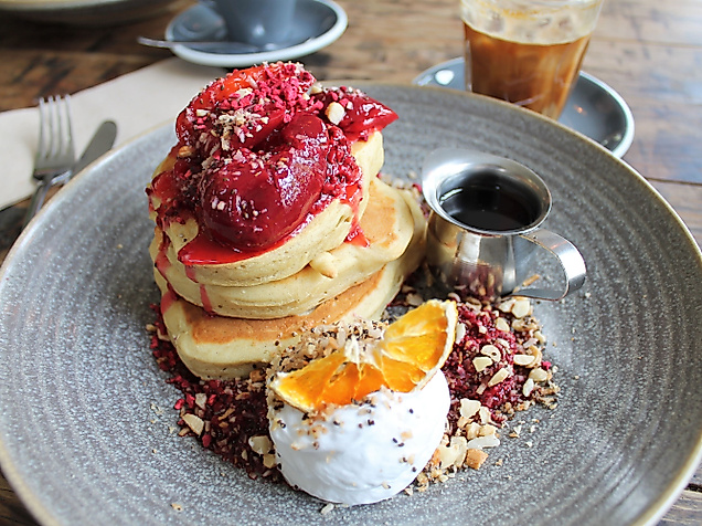 Pancake-Frühstück mit Beeren, Kokoscreme, Orangenscheibe und Ahornsirup auf grauem Teller angerichtet