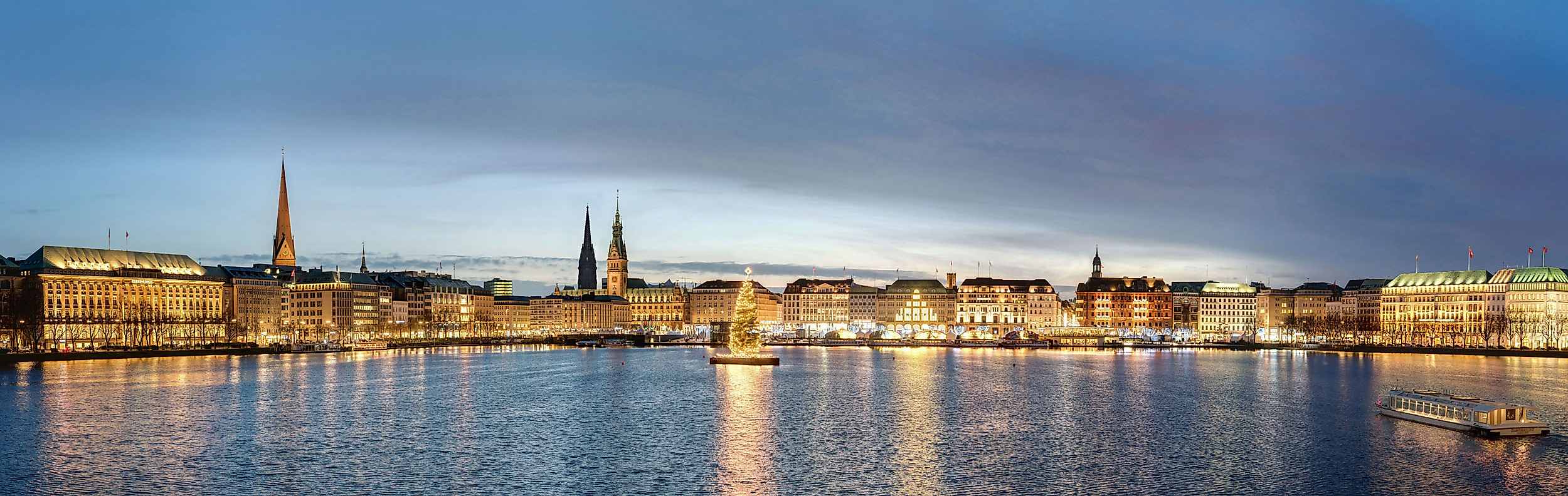 Abendstimmung an der Binnenalster mit Weihnachtsbaum, festlich beleuchteten Gebäuden und Ausflugsboot in Hamburg.