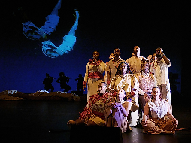 A group of performers in beige and red costumes sit and stand in formation on stage. In the background, silhouettes of musicians and a projection of a floating body are visible.