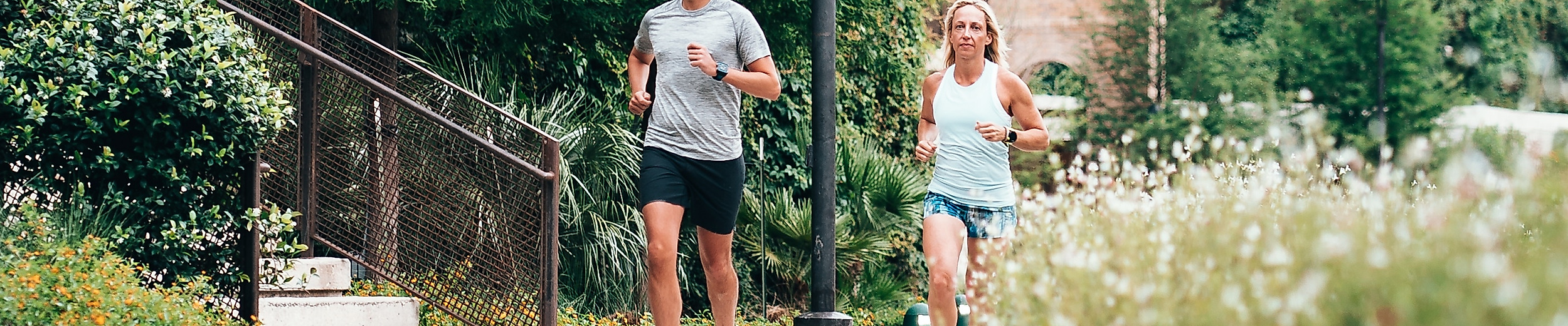 Zwei Jogger:innen laufen auf einem Weg durch eine grüne Stadtlandschaft mit Backsteinfassade im Hintergrund.
