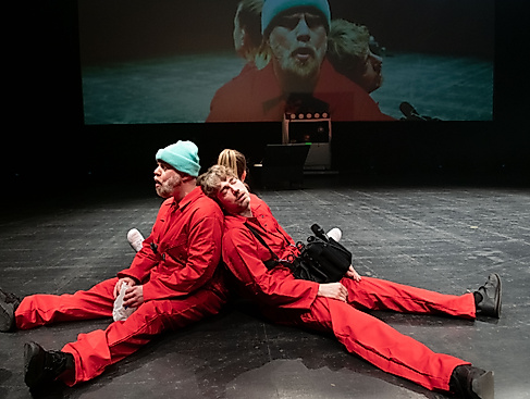 Drei Performer*innen in roten Overalls sitzen Rücken an Rücken auf einer Bühne und haben die Augen geschlossen. Hinter ihnen ist eine Nahaufnahme von ihnen auf die Wand projiziert.