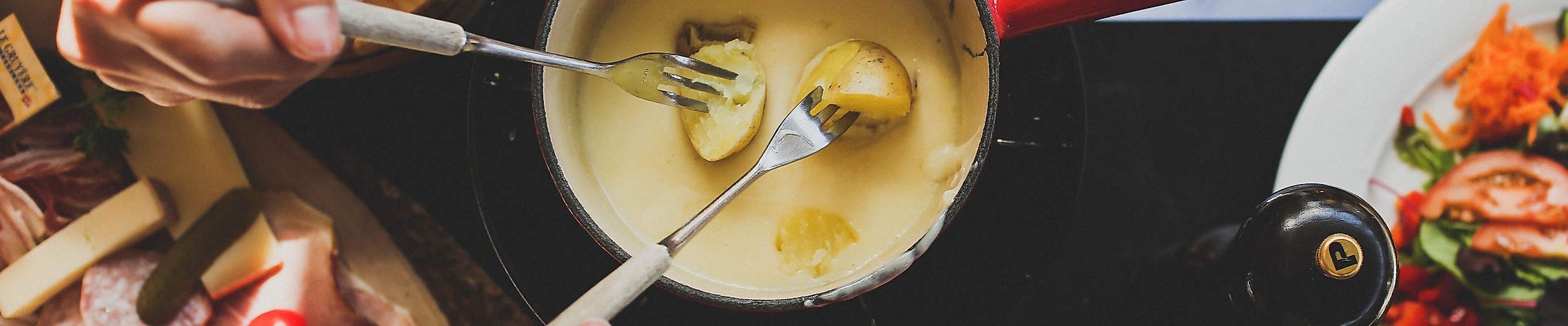Geselliges Käsefondue mit Brot, Kartoffeln und Beilagen auf gedecktem Tisch, zwei Personen tauchen ein