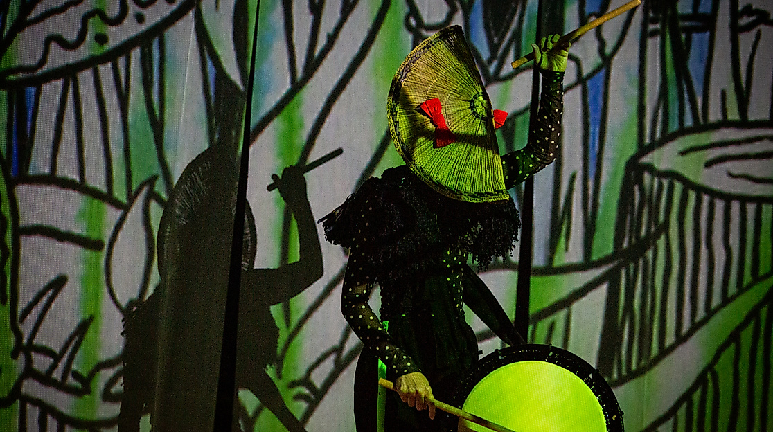 Eine Person trommelt vor einer in grün-schwarzen Symbolen angestrahlte Wand. Die eine Hand mit einem Drumstick in der Höhe, die andere an der Trommel. Auf dem Kopf sitzt eine fächerartige Kopfbedeckung.