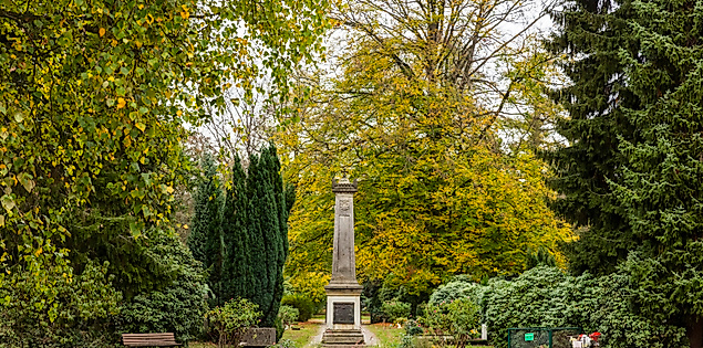 Denkmal im Ohlsdorfer Friedhof, umgeben von Bäumen, Bänken und gepflegter Bepflanzung in ruhiger Parklandschaft.