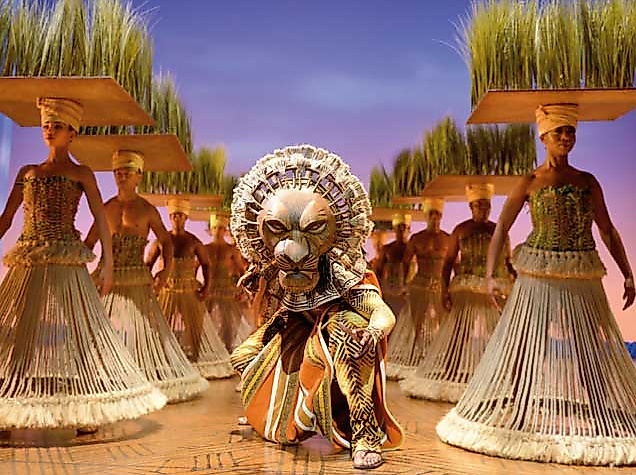 Szene aus dem Musical „Der König der Löwen“ in Hamburg: Im Zentrum ein kunstvoll kostümierter Löwencharakter, umgeben von Tänzer:innen mit großen Kopfbedeckungen in Form von Gräsern. Die Bühne ist in warmes Licht getaucht, der Hintergrund zeigt einen violetten Himmel. Ausdrucksstarke Darstellung afrikanischer Kultur und Tierwelt – ein unvergessliches Erlebnis, das mit der Hamburg CARD vergünstigt besucht werden kann.